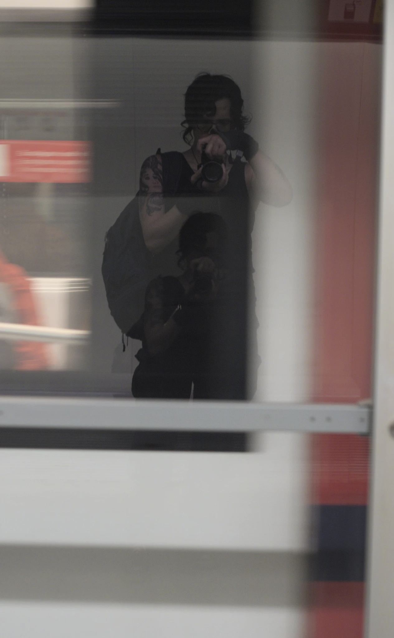 A double reflection shot of the photographer, who is made out in unusually clear detail here, on a glass reflection with a fast moving (blurry) red and white train behind the glass pane. The photographer is wearing all black, has a visible medusa tattoo, medium length hair, a backpack, and is holding the camera directly in front of her. You might say she's something of a very very soft futch in terms of style. Few details of her face can be made out, as the camera is obscuring at least part of it. Within the black mass that is her clothing, a second, smaller reflection of her appears.