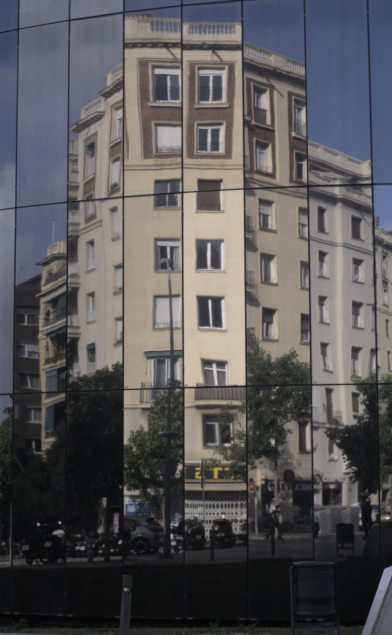 A photograph of the reflection of a building in the glass panes of another building. The glass building has a curved surface made up of tall, straight, flat glass planes linked together at an angle to form the curve. This has the visual effect of 'breaking up' any large reflection that would take multiple panels; some panels have a brighter reflection, some are darker or almost cloudy, and due to the angle difference some pieces of the reflected building are jutting up uncomfortably against sky or different parts of the building they don't encounter in real life; it's as if they took the 8 story slate brown building with dark brown walls on the top two floors and cut out various close-but-not-contiguous columnar regions, then re-arranged them into a somewhat flat surface. Note that the distortion/cutout effect is _only_ vertical; any given column of glass panes has the same angle, so the reflection going up from any point is continuous. The building being reflected, in reality, is at an angle here (it's a building on a corner, so it has at least two faces exposed to the street), but it's hard to tell. Various sections of trees are visible in the panels as well; some sections are brighter and others are dark. They have the same cut at random effect as the building itself.